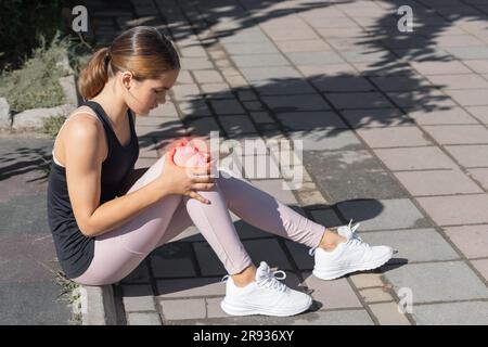 Das Mädchen hält ein verletztes Kniegelenk in den Händen. Verletzungen beim Laufen im Freien. Stockfoto