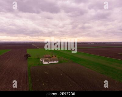 Luftflug über Winterlandwirtschaftsfelder mit verlassener Ranch bei Sonnenuntergang. Vojvodina, Serbien, Europa. Stockfoto