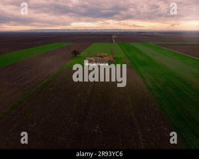 Luftflug über Winterlandwirtschaftsfelder mit verlassener Ranch bei Sonnenuntergang. Vojvodina, Serbien, Europa. Stockfoto