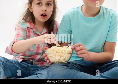 Kinder, die mit einer Schüssel Popcorn zurechtkommen, sehen fasziniert aus, während sie sich einen Film ansehen, isoliert auf weißem Studiohintergrund Stockfoto