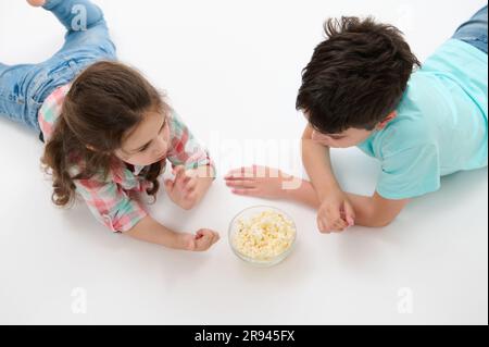 Draufsicht, junger Junge und süßes kleines Mädchen, Bruder und Schwester essen Popcorn, liegen auf weißem, isoliertem Studiohintergrund. Stockfoto