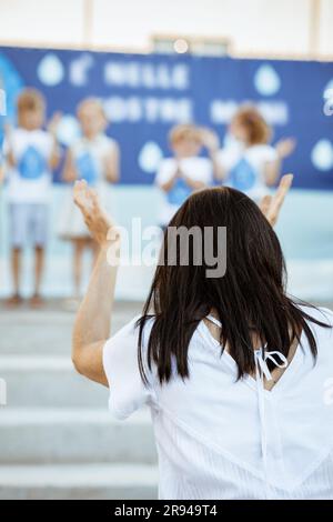 Der Rücken einer Frau im Vordergrund klatscht in die Hände und sieht sich eine Gruppe von Kindern auf der Bühne im Freien auf verschwommenem Hintergrund an Stockfoto