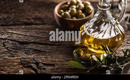 Eine Glasflasche Olivenöl und Blätter mit Oliven sind auf einem Holztisch entworfen. Stockfoto
