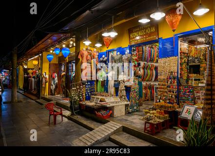 Kleidung und Souvenirläden mit ihren Waren bei Nacht entlang einer Straße in der Altstadt von Hoi an, Vietnam. Stockfoto