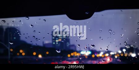 Regenschauer auf der Windschutzscheibe oder dem Autofenster und unscharfe Straße im Hintergrund. Fahren in der Regenzeit. Regentropfen auf dem Spiegel. Die Straße am Abend Stockfoto