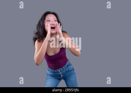 Junge asiatische Frau in einer schreienden Geste mit Händen in der Nähe ihres Mundes. Isoliert auf grauem Hintergrund. Stockfoto