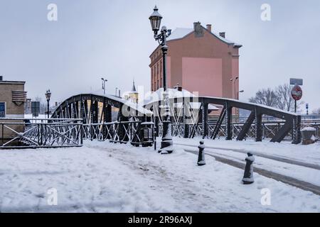 Klodzko, Polen - Januar 2023: Kleine Brücke mit schwarzen Metallbauteilen über einem kleinen Fluss voller Schnee im Winter Stockfoto