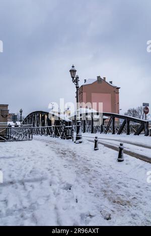 Klodzko, Polen - Januar 2023: Kleine Brücke mit schwarzen Metallbauteilen über einem kleinen Fluss voller Schnee im Winter Stockfoto