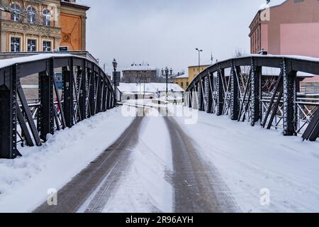 Klodzko, Polen - Januar 2023: Kleine Brücke mit schwarzen Metallbauteilen über einem kleinen Fluss voller Schnee im Winter Stockfoto