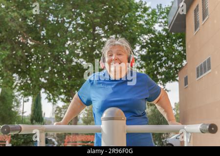 Aktive ältere Frau in Sportbekleidung trainiert auf einem Fitnessgerät im Park, während sie Musik über ihre Kopfhörer hört und die Kamera anlächelt Stockfoto