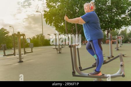 Aktive, fettleibige ältere Frau in Sportbekleidung trainiert auf einem Parkautomaten, während sie Musik über ihre Kopfhörer hört Stockfoto