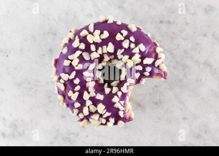 Donut mit lila Glasur, garniert mit einem kleinen weißen Schokoladenkrümel, wobei ein Biss fehlt. Stockfoto