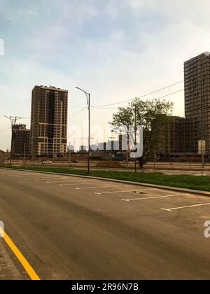 Stadtpanorama. Hohe Glashäuser stehen entlang einer Asphaltstraße mit gelben Streifen. In der Nähe von Lampenmasten, um die Straße zu beleuchten, und grüne Bäume mit Kronen. Stockfoto