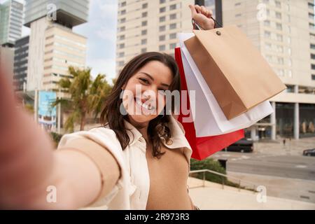 Ein weißes Mädchen nimmt ein fröhliches Selfie und hält Einkaufstüten mit erhobenem Arm. Stockfoto