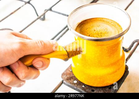 In seiner Hand ein gelber Türke mit Kaffee auf dem Herd. Stockfoto