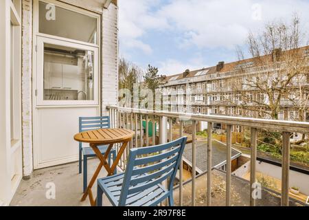 Ein Balkon mit zwei blauen Stühlen und einem Tisch auf der linken Seite, mit Blick auf ein Stadtviertel im Hintergrund Stockfoto