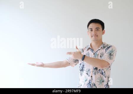 Ein junger asiatischer Mann, der ein rosa Hemd mit tropischen Mustern und eine Smartwatch am linken Handgelenk trägt, zeigt auf die rechte Seite seines offenen Arms Stockfoto