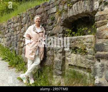 Eine grauhaarige Frau steht gegen eine Steinmauer und lächelt in die Kamera Stockfoto