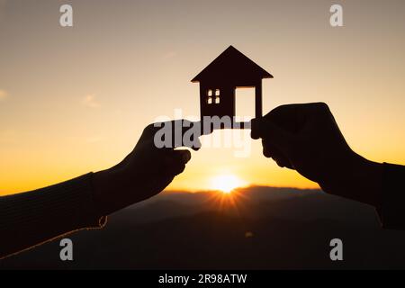Silhouette von männlichen und weiblichen Händen, die Modellhaus bei Sonnenuntergang halten Konzept des Kaufs von Häusern, Immobilien. Stockfoto