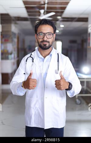 Porträt eines jungen männlichen Arztes mit Daumen nach oben. Glücklicher Seniorarzt im weißen Labormantel auf Krankenhauskorridorhintergrund. Vertrauen. Stockfoto