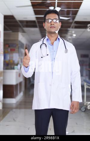 Porträt eines jungen männlichen Arztes mit Daumen nach oben. Glücklicher Seniorarzt im weißen Labormantel auf Krankenhauskorridorhintergrund. Vertrauen. Stockfoto