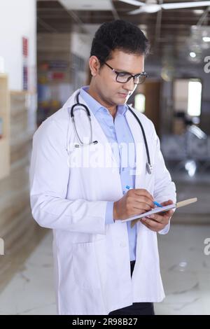 Porträt eines männlichen Oberarztes, der Notizen in Not Pad schreibt. Glücklicher indischer Arzt lächelnd, mit weißem Mantel im Krankenhausflur stehend. Stockfoto