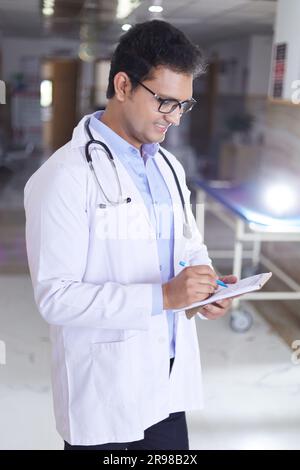Porträt eines männlichen Oberarztes, der Notizen in Not Pad schreibt. Glücklicher indischer Arzt lächelnd, mit weißem Mantel im Krankenhausflur stehend. Stockfoto