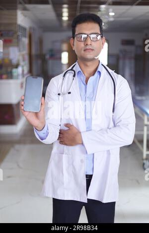 Porträt eines klugen jungen männlichen Arztes im weißen Labormantel mit Bildschirm des Mobiltelefons in der Hand. Doktor im Mantel lächelt auf Krankenhauskorridor Hintergrund. Stockfoto