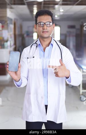 Porträt eines klugen jungen männlichen Arztes im weißen Labormantel mit Bildschirm des Mobiltelefons in der Hand. Doktor im Mantel lächelt auf Krankenhauskorridor Hintergrund. Stockfoto