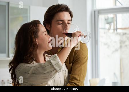 Lächelnde junge Frau, die ihren Freund Rotwein aus ihrem Glas trinken lässt Stockfoto