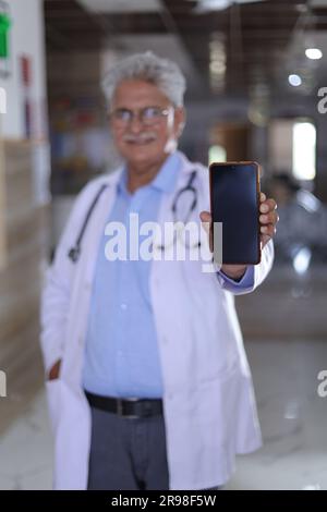 Porträt eines klugen Oberarztes im weißen Labormantel mit Bildschirm des Mobiltelefons in der Hand. Doktor im Mantel lächelt auf Krankenhauskorridor Hintergrund. Stockfoto