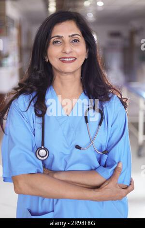 Porträt einer Ärztin mit überkreuzten Armen. Glückliche Krankenschwester in blauer Uniform, die im Krankenhauskorridorhintergrund steht. Vertrauen Stockfoto