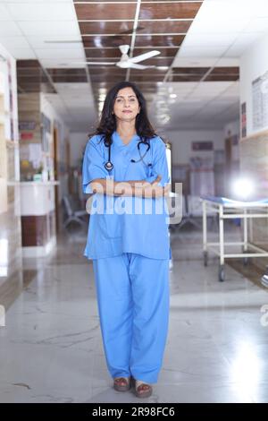 Porträt einer Ärztin mit überkreuzten Armen. Glückliche Krankenschwester in blauer Uniform, die im Krankenhauskorridorhintergrund steht. Vertrauen Stockfoto