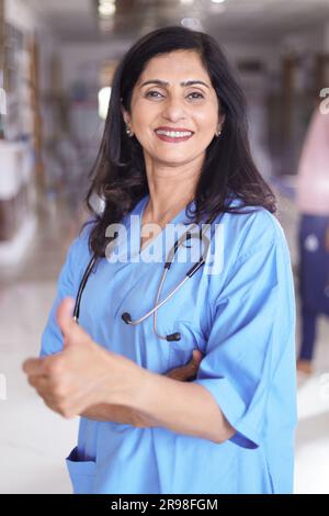 Porträt einer Ärztin mit Daumen nach oben. Glückliche Krankenschwester in blauer Uniform, die im Krankenhauskorridorhintergrund steht. Stockfoto