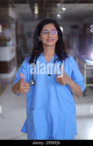 Porträt einer Ärztin mit Daumen nach oben. Glückliche Krankenschwester in blauer Uniform, die im Krankenhauskorridorhintergrund steht. Stockfoto