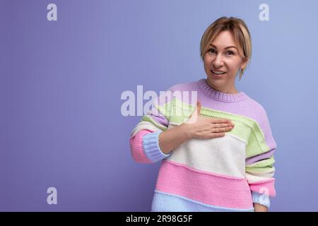 Lächelnde blonde junge Frau, die Hand an Seite auf lila Hintergrund zeigt Stockfoto