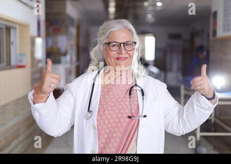 Senior Medical Professional Standporträt im Flur des Krankenhauses. Indische Ärztin, die mit beiden Händen die Daumen hoch hält. Stockfoto