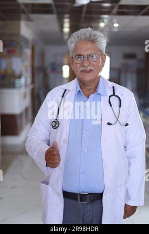 Porträt eines lächelnden männlichen Arztes im Laborkittel mit Daumen nach oben. Glücklicher Seniorarzt im weißen Mantel lächelnd auf Krankenhauskorridorhintergrund. Selbstbewusst. Stockfoto