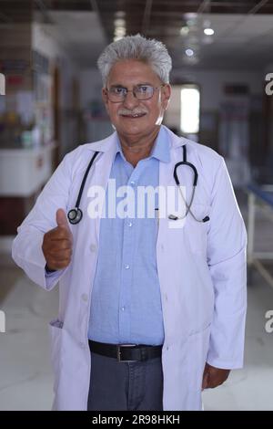 Porträt eines lächelnden männlichen Arztes im Laborkittel mit Daumen nach oben. Glücklicher Seniorarzt im weißen Mantel lächelnd auf Krankenhauskorridorhintergrund. Selbstbewusst. Stockfoto