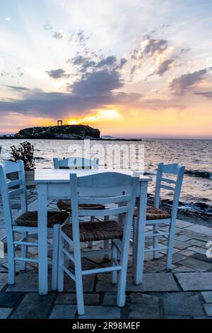 Der Sonnenuntergang in Richtung Portara in Chora auf der Insel Naxos, Griechenland, mit einem Tavernentisch und Stühlen im Vordergrund. Stockfoto