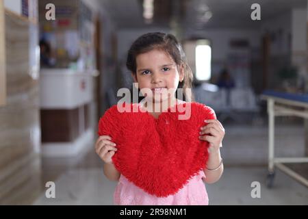 Kleines Mädchen Kind zeigt rot gefärbtes herzförmiges Kissen, steht im Krankenhausflur, zeigt Zuneigung, Dankbarkeit, Liebe, Patient geheilt. Stockfoto