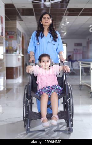 Kleines Mädchen Kind, das auf einem Rollstuhl sitzt und eine Krankenschwester sich um sie kümmert, beide stehen im Krankenhausflur, Verletzung, körperliche Behinderung. Stockfoto