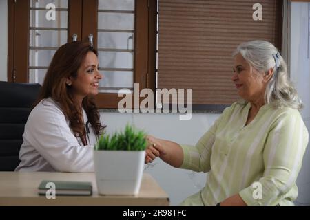 Junger Arzt, der mit Seniorenpatienten im Wohnzimmer des Pflegeheims sitzt. Älterer Patient mit ausführlichem Gespräch und Händeschütteln. Stockfoto