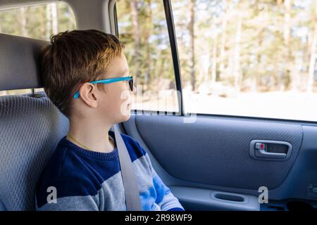 Ein weißer Junge im Schulalter fährt auf dem Rücksitz eines Autos und schaut aus dem Fenster. Das Kind legt den Sicherheitsgurt an und reist mit seinem Kind Stockfoto