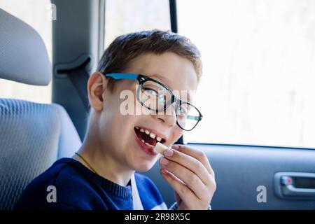 Ein glücklicher Weißer im Schulalter fährt auf dem Rücksitz eines Autos und isst Süßigkeiten. Ein Schuljunge mit Brille. Das Kind ist angeschnallt und er Stockfoto