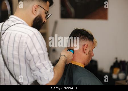 Bärtiger Barbier schneidet Haare von männlichen Kunden. Friseurin, der im Friseursalon arbeitet. Stockfoto