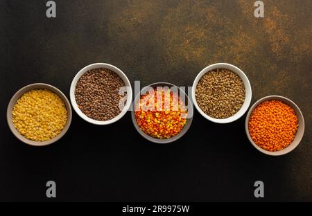 Mehrfarbige Linsen in Schüsseln auf braunem Hintergrund, gelb und braun, grüne und orangefarbene Linsen, gesunde Hülsenfrüchte, Draufsicht, Kopierbereich Stockfoto