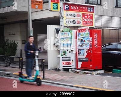 TOKIO, JAPAN - 9. Juni 2023: Ein Fahrer eines gemieteten E-Rollers fährt einen Hügel hinunter, vorbei an zwei Verkaufsautomaten auf einer Straße vor dem Parkplatz durch eine apa Stockfoto