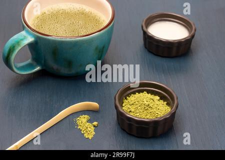 Grünes Matcha-Pulver und Messlöffel auf dem Tisch. Milch in der Schüssel. Matcha-Tee im Becher. Blauer Hintergrund Stockfoto