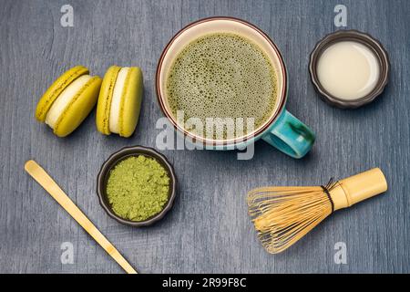 Messlöffel und grünes Matcha-Pulver. Milch in der Schüssel. Makkaron-Kekse und Matcha-Tee im Becher. Flach verlegt. Blauer Hintergrund Stockfoto